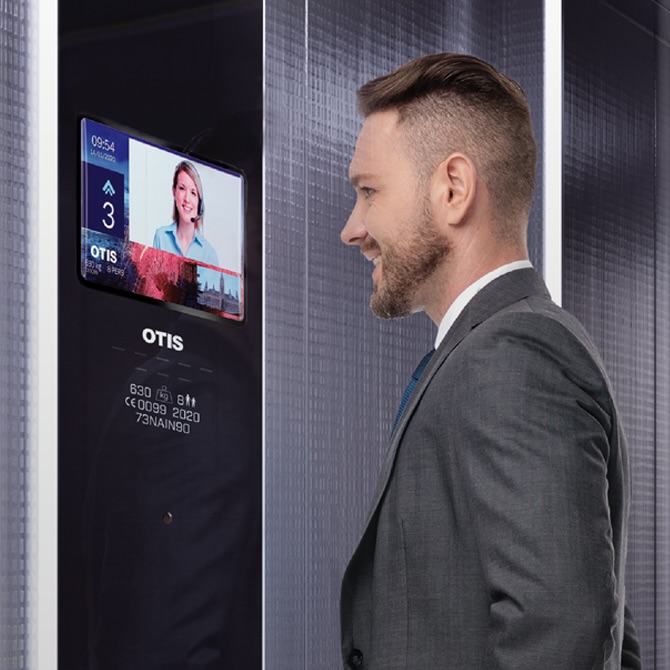 A man stands in a lift looking a digital screen in the cab, video-chatting with an Otisline agent.